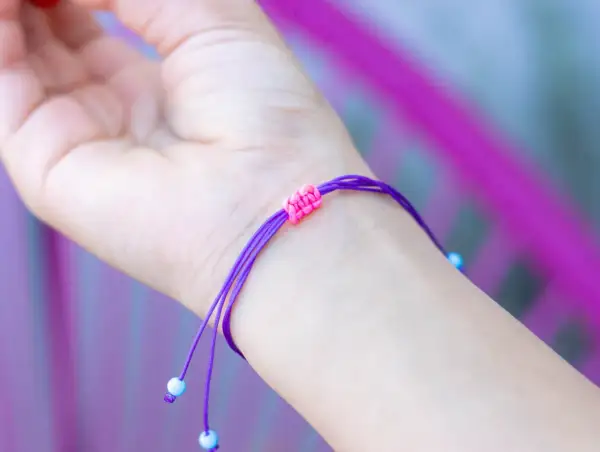 Eine Hand trägt ein lila Armband mit einem doppelten Schiebeknoten. Kleine blaue Perlen zieren die Enden.